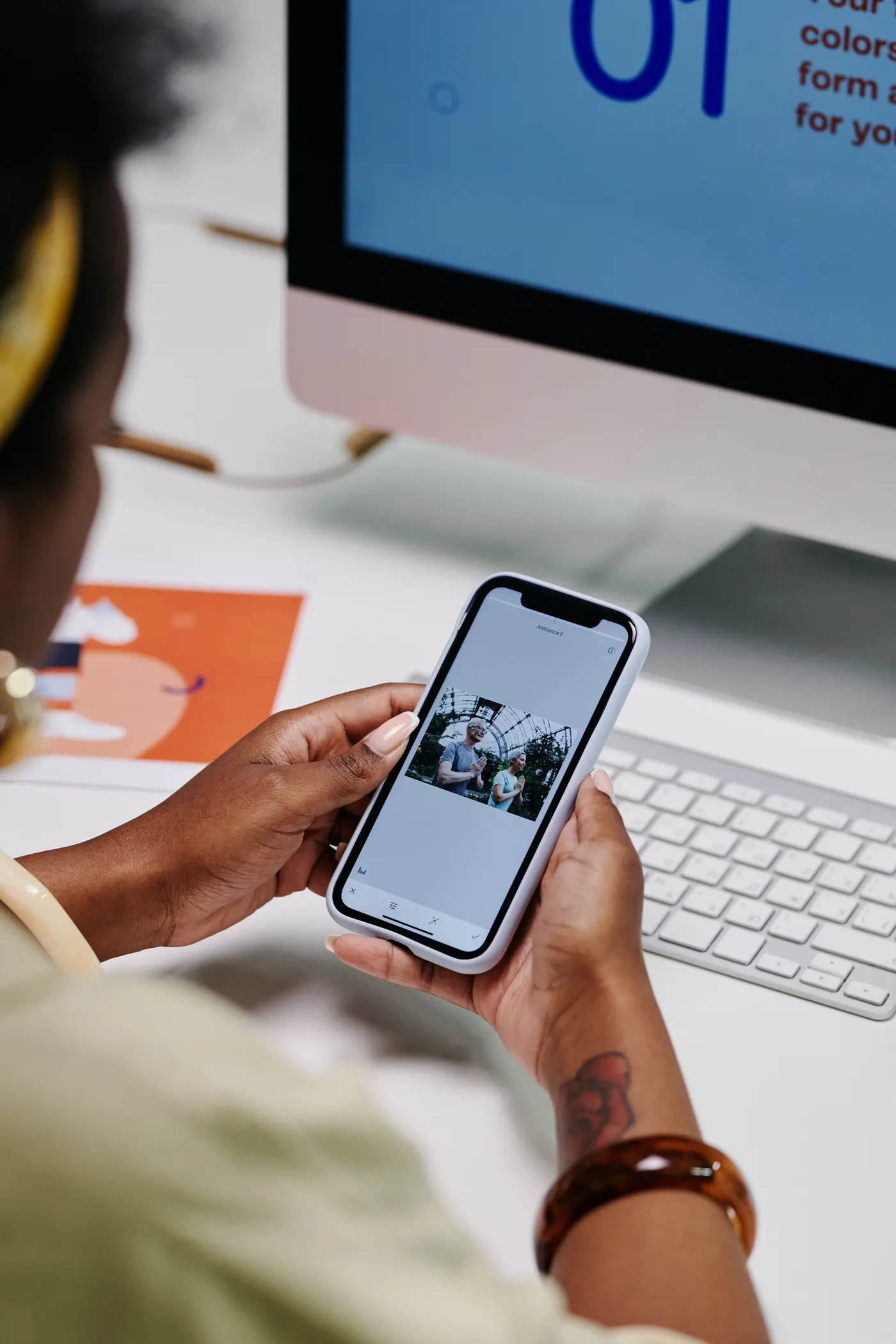 Naima reviewing brand work on a phone beside an iMac showing a colour-system layout